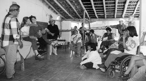 Trabajando con participantes del curso en PROJIMO, Jean Anne mantiene a Xochi entre sus piernas para explorar su rango de movimiento, cómo mejorar su posición y el tipo de asiento adaptativo, cuñas y soportes que la ayudarán a sentarse lo mejor que pueda.