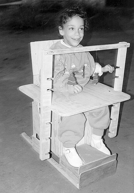 The seat includes a tray, a bar with hanging toys to stimulate eye-hand control, and a cardboard foot-separator.