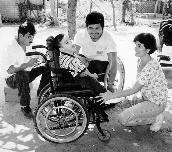 Gabriel and the mother of a boy with spastic cerebral palsy try the child in his new wheelchair, specially adapted with an adjustable head-rest, seat-belt, and shoulder straps. Meanwhile Juan takes notes for additional adjustments that are needed for better function.